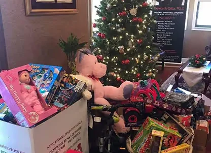 Collection of toys including a large pink teddy bear and boxed dolls piled next to a decorated Christmas tree in a room.