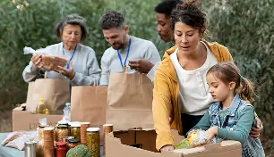 People packing food items into boxes outdoors, including a child and three adults organizing jars and bags.