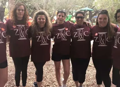 Group of six women standing outdoors with arms around each other, all wearing matching maroon shirts with 'ZINC' printed on them.