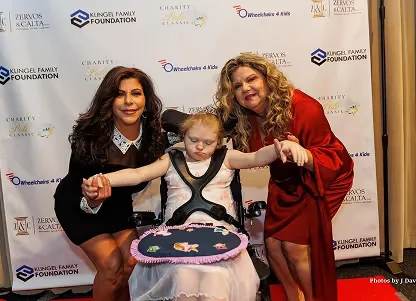 Two women holding hands of a young girl in a wheelchair at a charity event with branded backdrop.