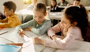 Two young girls sitting at a desk in a classroom, writing in notebooks, with other children in the background.