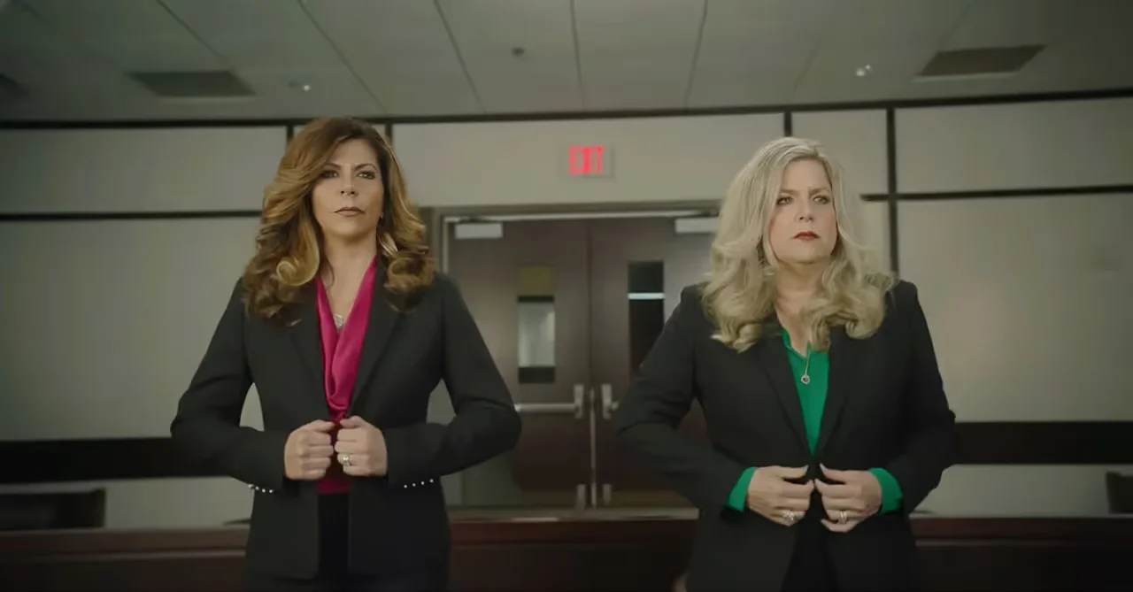 Two women in black suits buttoning their jackets in a courtroom, one with a magenta blouse and the other with a green blouse.