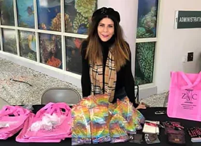 Woman wearing a black beret and scarf standing behind a table with colorful bags and pink reusable bags in an indoor setting.