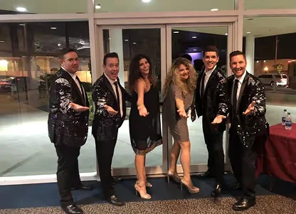 Group of five men in black suits with sequined jackets and two women in elegant dresses posing indoors with arms extended forward.